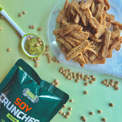 Close up of wasabi flavoured soy crunches surrounded by scattered soybeans and a spoonful of wasabi on a light green background. 