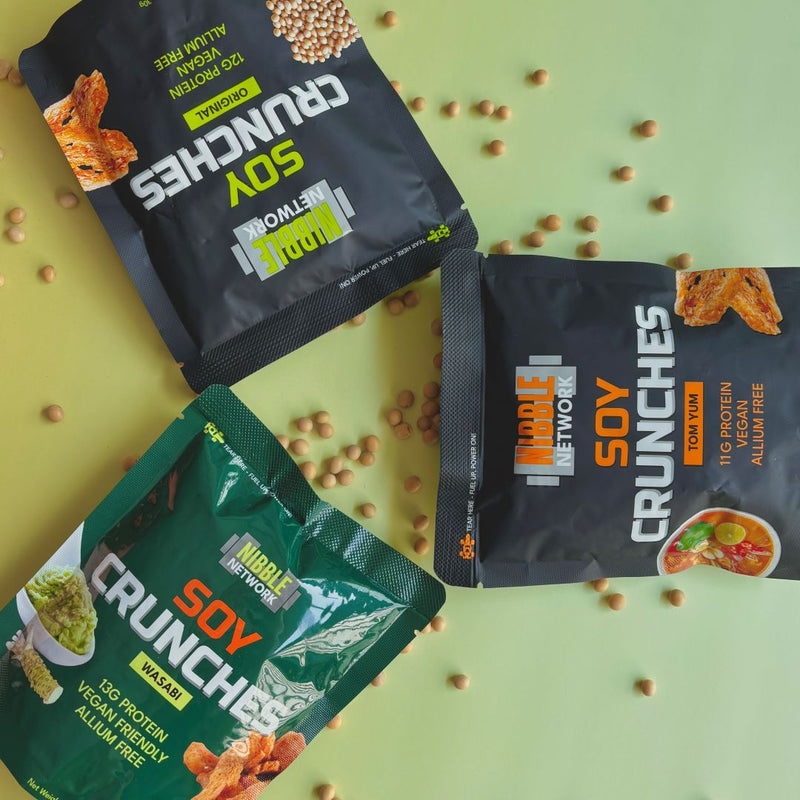Three packages of Nibble Network Soy Crunches on a green background with scattered soybeans.