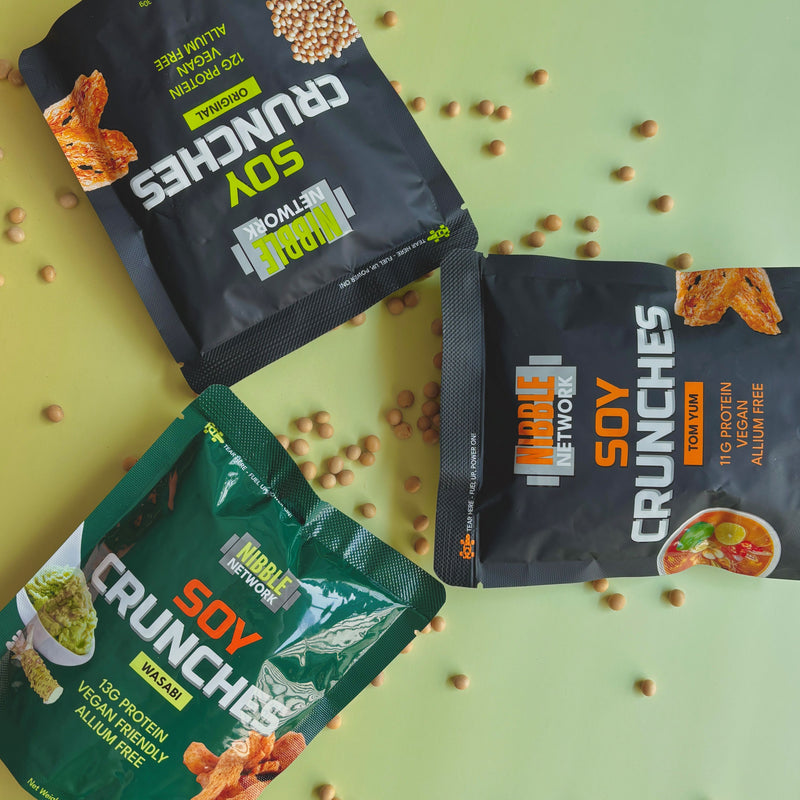 Three packages of Nibble Network Soy Crunches on a green background with scattered soybeans.